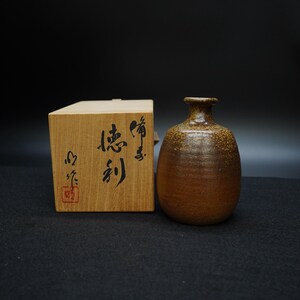 Puede incluir: Una botella de sake de cerámica marrón con cuello estrecho y cuerpo redondeado, junto a una caja de madera. La botella tiene un acabado texturizado y moteado. La caja de madera tiene caligrafía japonesa.