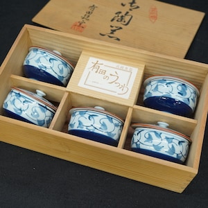 以下が含まれることがあります： 青と白の蔓模様が描かれた蓋付きの陶器製ボウル5個セット。長方形の木箱に収められており、中央には「有田のうつわ」と書かれた白いラベルが貼られています。木製の蓋が近くに置かれています。