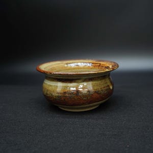 May include: A small, handcrafted ceramic bowl with a wide rim. The bowl features a color palette of brown, tan, and cream, with visible brushstrokes. The interior is a lighter shade, while the exterior has darker bands. The bowl is likely for decorative or functional use.