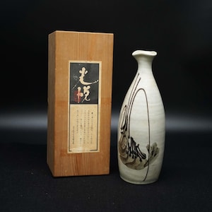May include: A white ceramic sake bottle with a narrow neck and a brown abstract design. The bottle is next to a wooden box with Japanese text on a label. The box is made of light-colored wood.