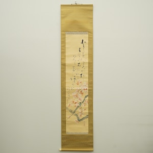 Puede incluir: Pintura japonesa vertical en rollo con borde de brocado dorado. La obra presenta una rama de flores de cerezo con un pequeño pájaro y caligrafía en tinta negra. El rollo está colgado contra una pared neutra.