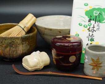 Op de afbeelding: Een Japanse theeceremonieset bestaande uit een bruine gespikkelde keramische kom met een bamboe garde, een lichte keramische kom, een donkerrode gelakte container met gouden cirkelpatronen, een kleine witte keramische houder, een bamboe lepel en een witte doos.