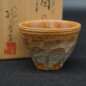 以下が含まれることがあります： 手びねりの陶器製ティーカップ。質感のあるアースカラーの茶色と灰色の釉薬が施されています。カップは広い開口部と先細りの底が特徴です。黒い日本語の書道と赤い印章が押された明るい色の木箱の前に置かれています。