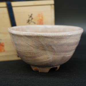 May include: A ceramic tea bowl with a light beige and soft purple marbled glaze featuring a fine crackle finish. The bowl has a slightly irregular, hand-formed shape with a flared rim and a small footed base, positioned in front of a wooden box.