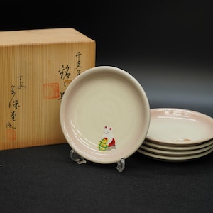 以下が含まれることがあります： 薄いピンク色の縁とクリーム色の中心部を持つ、4枚のセラミックプレートのセット。各プレートには、小さなカラフルなキャラクターのイラストが描かれています。背景には日本語のテキストが入った木箱が見えます。