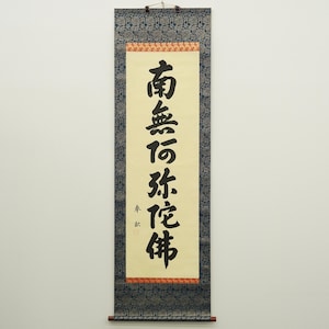 May include: A traditional Japanese scroll with black calligraphy on a cream-colored background. The text is framed by a red and gold border, with a dark blue patterned fabric at the top and bottom. A wooden rod is at the bottom.