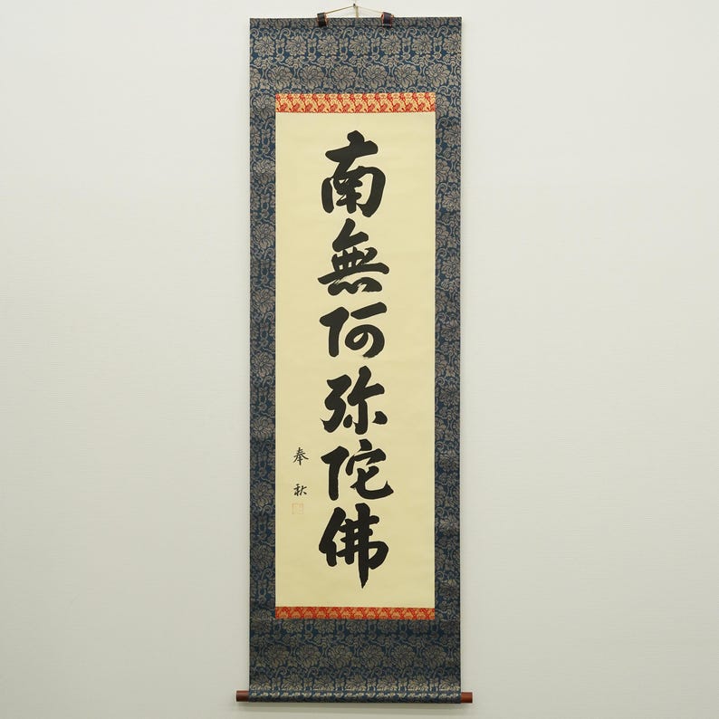 May include: A traditional Japanese scroll with black calligraphy on a cream-colored background. The text is framed by a red and gold border, with a dark blue patterned fabric at the top and bottom. A wooden rod is at the bottom.