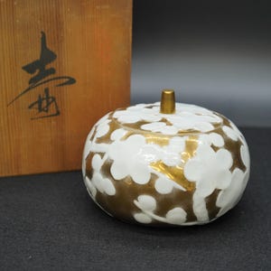 以下が含まれることがあります： 白いと金のセラミックジャー、蓋付き、花柄のデザイン。ジャーは金色のベースと蓋があり、白い花の模様があります。背景には、日本の文字が入った木箱があります。