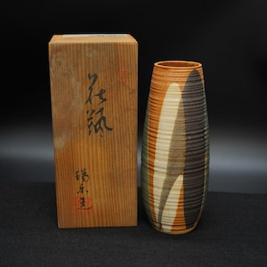 以下が含まれることがあります： 茶色、オレンジ、白、緑の色合いの、高さのある質感のあるセラミック花瓶。花瓶は、日本の書道が施された木箱の隣にあります。花瓶は、ユニークなリブのデザインです。