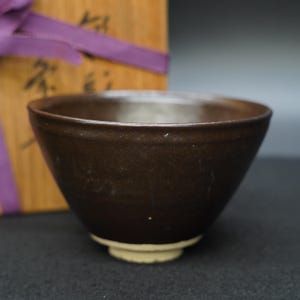 May include: A dark brown ceramic bowl with a slightly flared rim and a light beige base. The bowl has a smooth, matte finish. A wooden box tied with a purple ribbon is in the background.