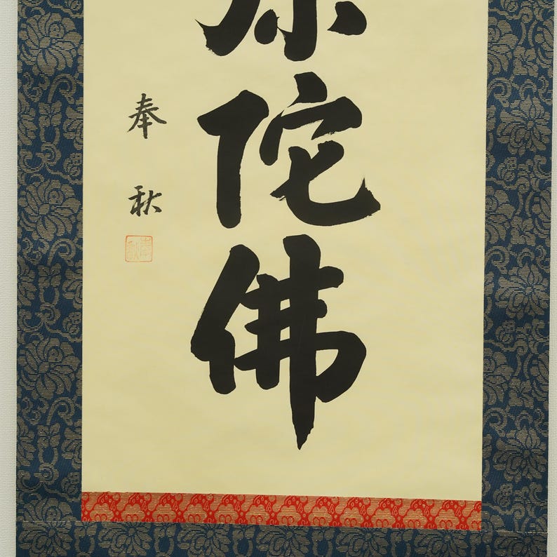 May include: A traditional Japanese scroll featuring calligraphy in black ink on a cream-colored background. The scroll includes several characters and symbols, with a red patterned border at the bottom and a blue floral patterned border on the sides.