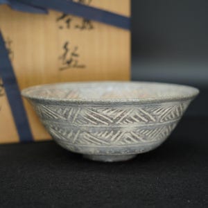 May include: A gray ceramic bowl featuring a textured, incised geometric pattern of repeating diagonal lines around the exterior. The bowl is positioned in front of a light-colored wooden box tied with a dark blue ribbon and marked with black ink text.