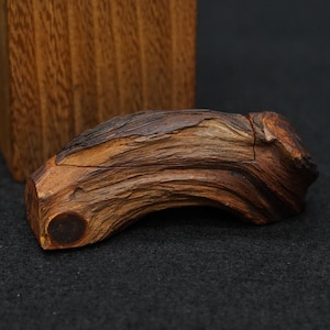 以下が含まれることがあります： 自然で有機的な形をした手作りの木箱。木箱は、木目と質感が目立つ濃い茶色の木材で作られています。蓋はわずかに湾曲しており、箱の端には小さな丸いディテールがあります。背景には木製のブロックが含まれています。