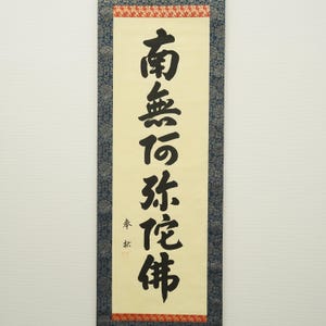 May include: A vertical scroll with black calligraphy on a cream-colored background. The text is in a traditional East Asian script. The scroll is framed with a blue patterned border and red accents.