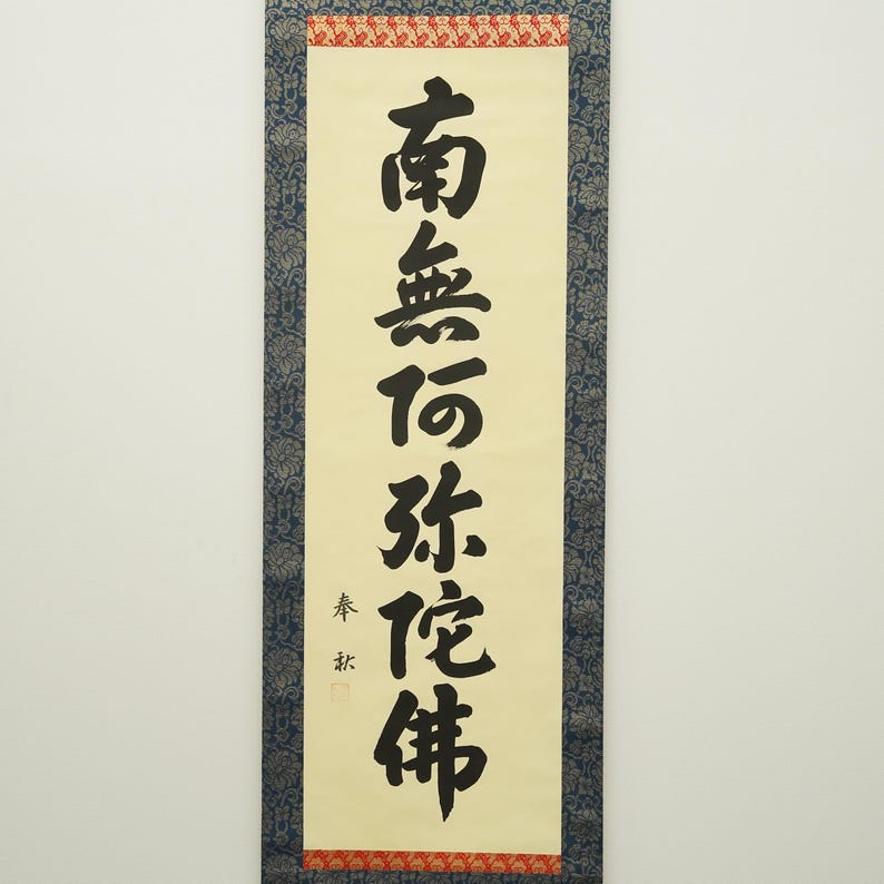 May include: A vertical scroll with black calligraphy on a cream-colored background. The text is in a traditional East Asian script. The scroll is framed with a blue patterned border and red accents.