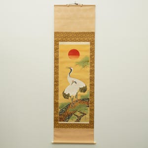 Peut inclure: Une peinture sur rouleau vertical représentant deux grues blanches aux pointes d'ailes noires perchées sur une branche d'arbre brune texturée. Au-dessus des oiseaux, un grand soleil rouge brille sur un fond jaune. Bordures dorées à motifs.