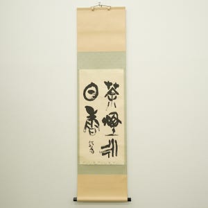 以下が含まれることがあります： 黒インクの書道が施された、縦長の日本の巻物。巻物はクリーム色で、薄緑色とタン色の縁取りがあります。書道にはいくつかの文字と円形の記号が含まれています。