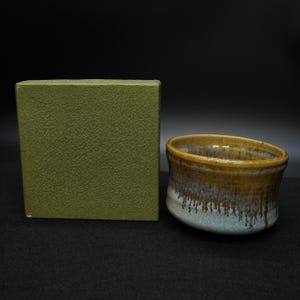 Peut inclure: Un bol en céramique fait à la main avec un glaçage unique. Le bol présente un dégradé de couleurs, notamment du marron, du blanc et du bleu. À côté du bol se trouve une boîte verte texturée, probablement pour offrir ou ranger. La conception du bol suggère qu'il pourrait être utilisé pour servir ou exposer.