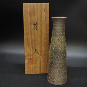 Puede incluir: Un jarrón de cerámica alto y texturizado con un degradado marrón y verde. El jarrón tiene un patrón de panal y está junto a una caja de madera con caracteres japoneses. La caja tiene un sello rojo.