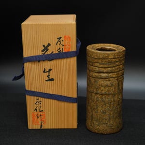 Op de afbeelding: Een cilindervormige, getextureerde, bruine keramische vaas met een cirkelvormige opening, naast een houten doos die met een blauw lint is vastgebonden. De doos heeft Japanse karakters en rode stempels. De vaas heeft horizontale ribbels.