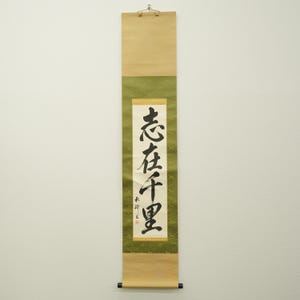 以下が含まれることがあります： 黒い書道が施された伝統的な日本の掛け軸。掛け軸は、上部と下部がベージュで、中央に緑色の模様の部分があります。書道は白い背景に配置されています。