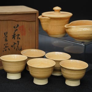 以下が含まれることがあります： 木箱、蓋付きの急須、盛り鉢、6つの小さな湯呑み茶碗がセットになった日本のティーセット。 ティーセットは薄いオレンジ色で、湯呑み茶碗には白い台座が付いています。 木箱には日本語の文字が書かれています。