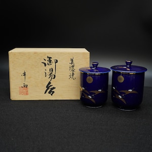 May include: Two cobalt blue ceramic tea cups with lids, featuring gold accents and a crane design. The cups are displayed with a wooden box that has Japanese text on it. The cups are likely part of a tea set.