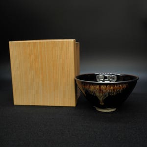May include: A black ceramic bowl with a brown and gold glaze, next to a light brown wooden box. The bowl has a wide rim and a flared shape. The glaze has a unique, flowing pattern.