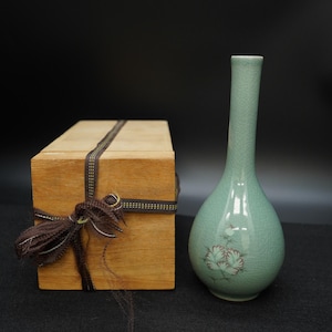 Può includere: Un vaso in ceramica color celadon con collo lungo e corpo bulboso, con un motivo floreale. Accanto, una scatola di legno legata con un nastro marrone. Il vaso ha una finitura a craquelé e la scatola è in legno chiaro.