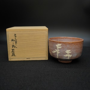 Puede incluir: Un cuenco de té de cerámica marrón rojizo con diseños abstractos blancos y una caja de madera. El cuenco tiene un borde ancho y una base con pie. La caja de madera tiene caracteres japoneses.