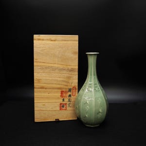 May include: A celadon ceramic vase with a long neck and a bulbous body, decorated with cranes and floral patterns. The vase is next to a wooden box with Asian characters and stamps.