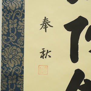 May include: A close-up of a traditional Japanese calligraphy artwork. The artwork features black ink characters on a cream-colored background, with a blue and gold patterned border on the left side. The calligraphy includes a red seal.