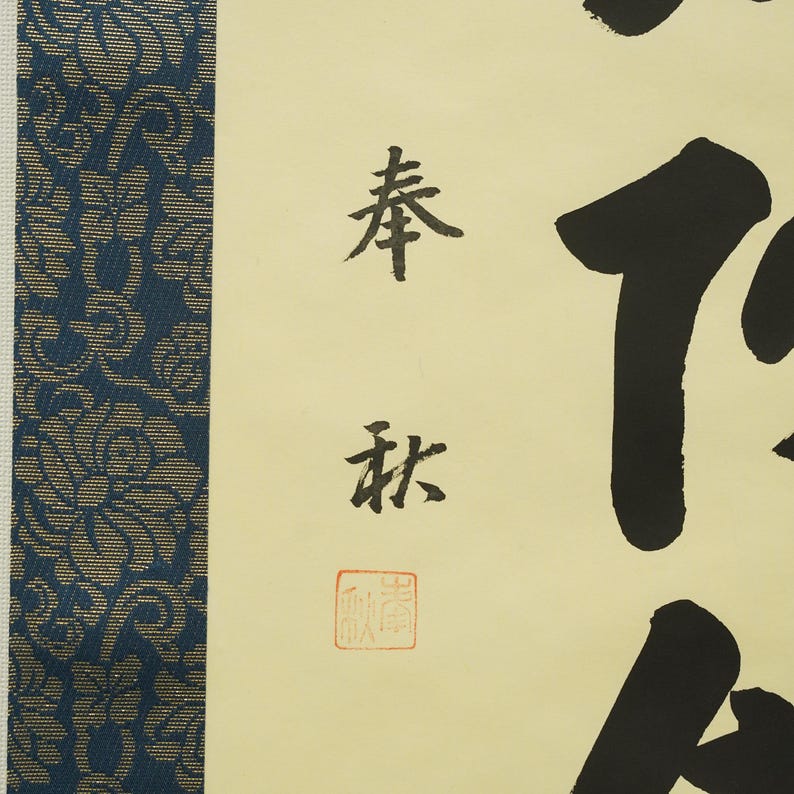 May include: A close-up of a traditional Japanese calligraphy artwork. The artwork features black ink characters on a cream-colored background, with a blue and gold patterned border on the left side. The calligraphy includes a red seal.