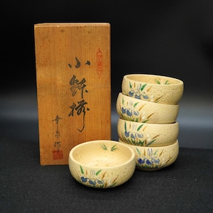 May include: A set of six beige ceramic bowls with blue and green floral designs, stacked and arranged next to a wooden box with Japanese calligraphy. The bowls have a textured surface.