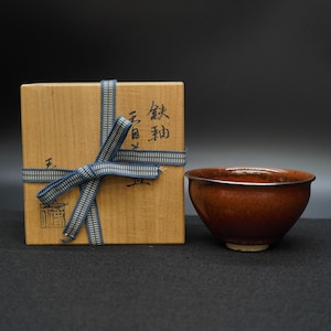 Pode incluir: Uma tigela de chá de cerâmica marrom com base branca está ao lado de uma caixa de madeira amarrada com uma fita azul. A caixa tem texto japonês. A tigela de chá tem um acabamento brilhante e uma cor marrom escura.