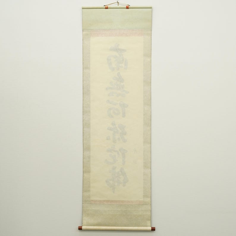 May include: A traditional Japanese scroll with calligraphy in black ink. The scroll features a light beige background with a green top and a brown bottom rod. The calligraphy is vertically oriented and centered on the scroll.