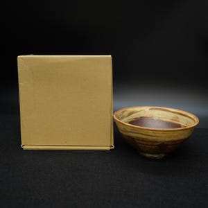 Puede incluir: Un cuenco de cerámica hecho a mano con un esmalte marrón y beige. El cuenco tiene forma redondeada y un interior marrón oscuro. A la izquierda del cuenco hay una caja de cartón lisa. El cuenco probablemente se utiliza para servir alimentos.