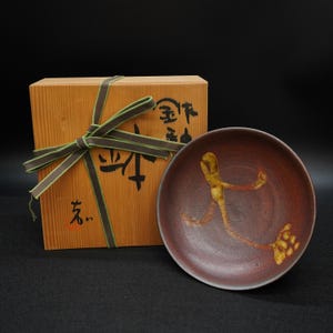 Puede incluir: Un plato de cerámica marrón con un diseño abstracto dorado y naranja. El plato está junto a una caja de madera atada con una cinta verde y marrón. La caja tiene caracteres japoneses.