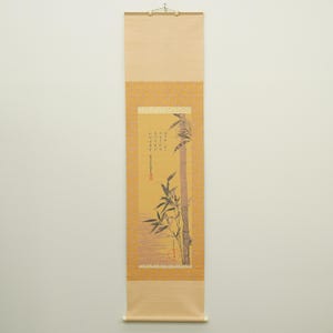 Pode incluir: Um rolo vertical para pendurar com uma pintura de caules de bambu sobre um fundo amarelo. A obra inclui caligrafia a tinta preta e um selo vermelho. O rolo é emoldurado por uma borda de tecido cor de mostarda com um padrão floral.