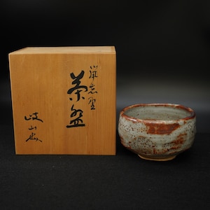 以下が含まれることがあります： 灰と茶色の斑点のある釉薬が施された陶器のティーボウルが、日本語の書道が施された木箱の隣にあります。 ボウルは丸みを帯びた形状で、表面はざらざらしています。