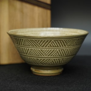 May include: A ceramic bowl with a footed base and a wide rim. The bowl features a repeating geometric pattern in a darker shade of green against a lighter background. The bowl is likely used for serving food.