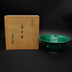 Op de afbeelding: Een groene keramische kom met een bloemmotief en een houten doos met Japanse karakters. De kom heeft een glanzende afwerking en staat naast de doos op een donkere ondergrond. De doos is gemaakt van lichtgekleurd hout.