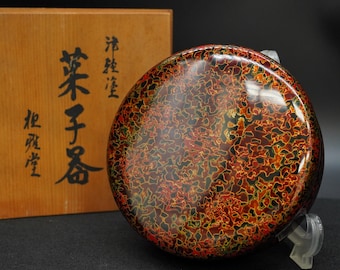May include: A round, lacquered container with a vibrant, patterned surface in shades of red, orange, and green. The container is displayed on a clear stand, with a wooden box featuring Japanese characters in the background.
