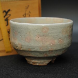 May include: A handcrafted ceramic tea bowl with a textured, light green exterior and subtle pink floral patterns. The bowl has a rounded shape with a footed base, suggesting traditional Japanese pottery. The background includes a wooden box.