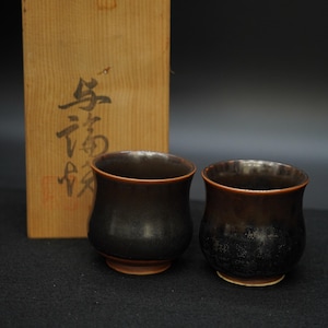 以下が含まれることがあります： 光沢のある仕上げと明るい茶色の縁と底を持つ、2つの濃い茶色のセラミックティーカップ。背景には、日本の書道が施された木箱があります。ティーカップは丸みを帯びた形をしています。