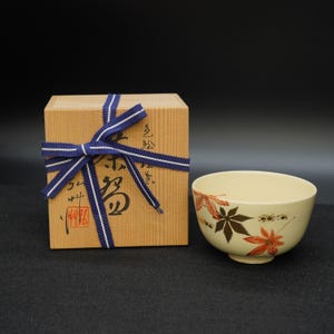 Puede incluir: Un cuenco de cerámica color crema con un diseño de hojas de arce pintadas en rojo y marrón. El cuenco está junto a una caja de madera atada con una cinta azul y blanca. La caja tiene texto japonés y un sello rojo.