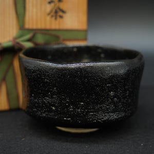 Könnte beinhalten: Eine schwarze Teeschale mit einer texturierten, unebenen Oberfläche und einem leicht unregelmäßigen Rand. Die Schale steht vor einer hellen Holzschachtel, die mit einem grünen Band gebunden ist und ein schwarzes japanisches Schriftzeichen trägt.