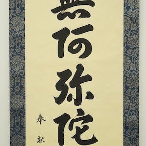 May include: A vertical scroll with black calligraphy on a cream-colored background. The calligraphy features large, bold characters. The scroll is framed by a dark blue floral patterned border. The text includes smaller characters at the bottom.