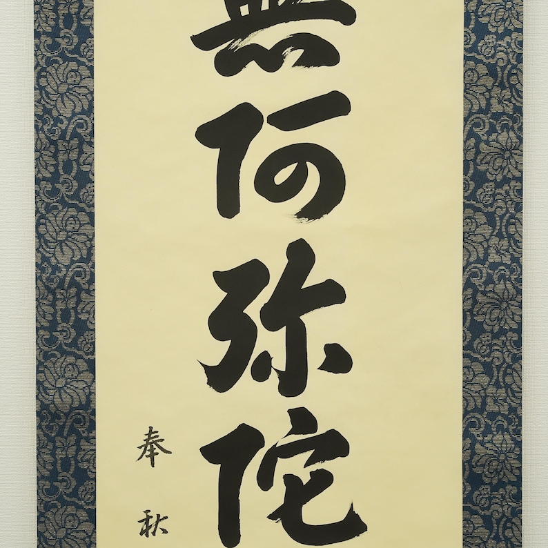 May include: A vertical scroll with black calligraphy on a cream-colored background. The calligraphy features large, bold characters. The scroll is framed by a dark blue floral patterned border. The text includes smaller characters at the bottom.