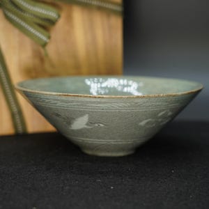 Puede incluir: Un cuenco de cerámica de color celadón con un diseño ancho y poco profundo. El cuenco presenta un patrón en relieve detallado con ilustraciones de grullas volando. El borde tiene un sutil acento marrón. El cuenco está sobre un fondo de madera borroso.